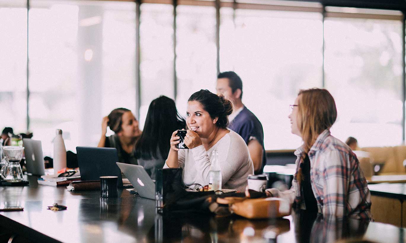 Two women sit and drink coffee while working on their laptops in a busy office setting.