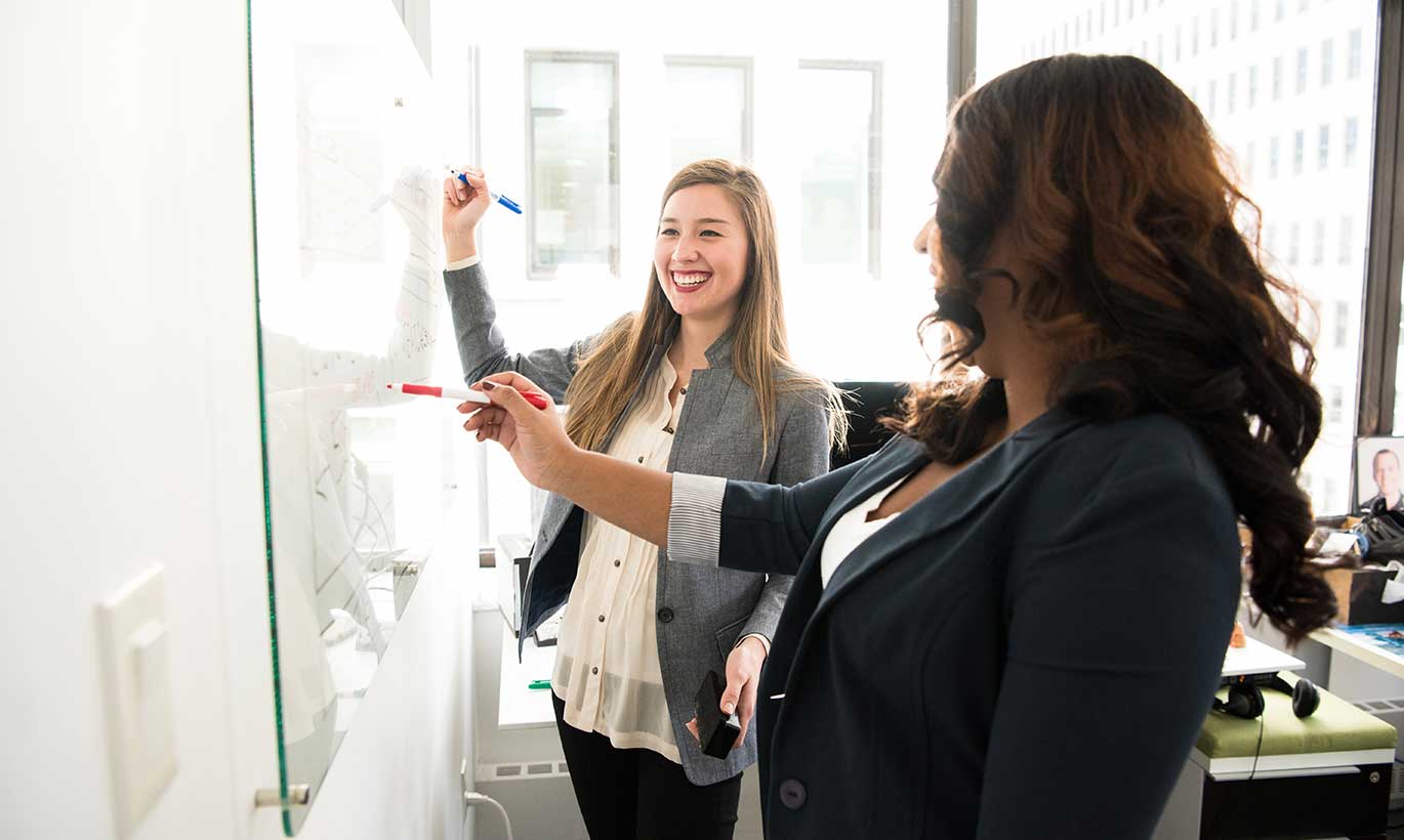 Two women in business clothing write on a whiteboard with Expo markers while talking and smiling.