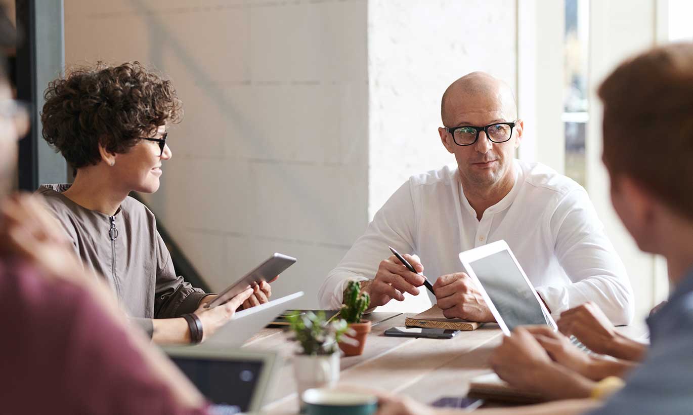 A man wearing glasses speaks to his three colleagues during a meeting while they all hold tablets
